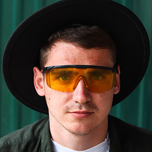 The Grant Getters A man wearing a wide-brimmed black hat and yellow-tinted protective glasses, looking like one of The Grant Getters, stares directly at the camera. He has short dark hair and a slightly serious expression. Grant Writing