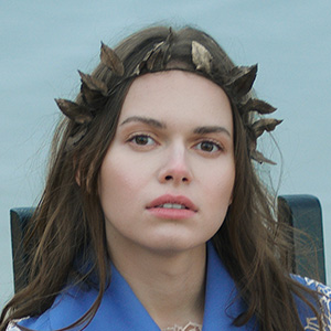 The Grant Getters A woman with long brown hair and a neutral expression wears a headband made of feathers. She has a blue garment draped over her shoulders, embodying the serene focus often seen in seasoned grant writers from The Grant Getters, and is outdoors with a cloudy sky in the background. Grant Writing